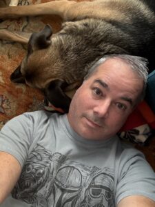 A man with gray hair lies on his back on a patterned rug, taking a selfie from above. He wears a gray t-shirt with a graphic design featuring what appears to be sci-fi helmets or masks. Behind him, a large dog with tan and black coloring is sprawled out on the same rug, lying belly-up with legs extended. The image captures a relaxed moment of companionship between person and pet.