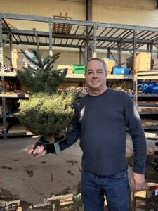 A man with gray hair stands in a warehouse, holding a small Christmas tree. He wears a dark blue textured sweater over jeans and has a slight smile. Behind him are industrial metal shelving units filled with storage bins and boxes. The concrete floor shows signs of wear, and the warehouse has high ceilings with exposed ductwork and beige walls.
