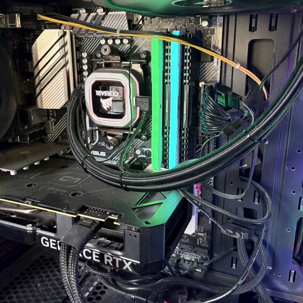 Interior view of a gaming PC build showing an ASUS motherboard with Corsair CPU cooler, green RGB RAM modules, GeForce RTX graphics card, and custom cable management with braided cables inside a case with RGB lighting.