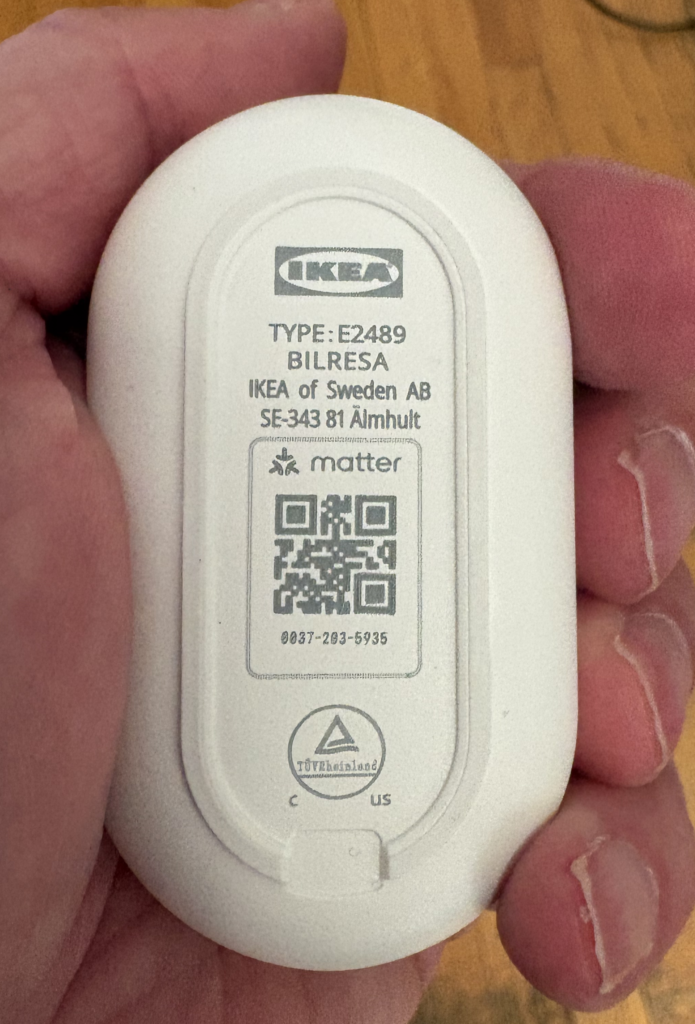 A hand holds a small white oval device labeled "IKEA BLE-ESA" with a QR code and product information printed on it.