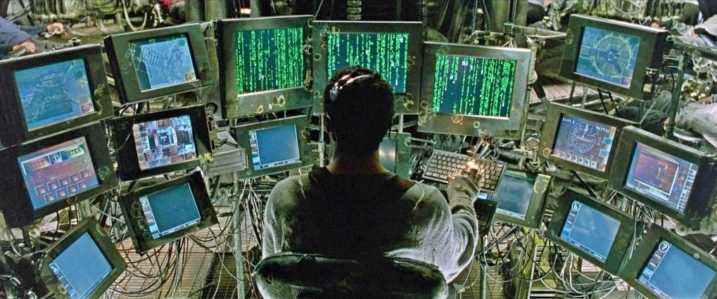 A person sits at a complex workstation surrounded by multiple computer monitors displaying green Matrix-style code and various digital interfaces, with cables and equipment scattered throughout the dimly lit space.