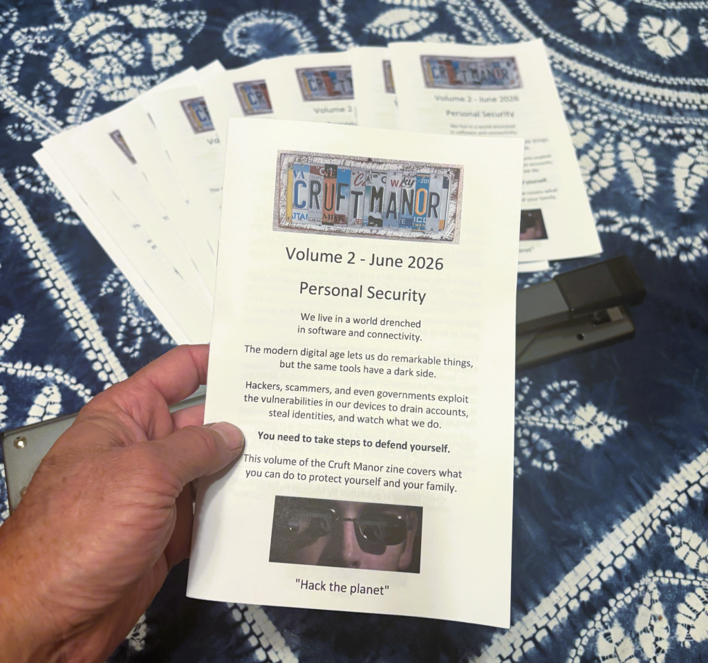 A hand holds multiple copies of the "Cruft Manor" zine, Volume 2 - June 2026, titled "Personal Security," featuring text about digital threats and the quote "Hack the planet," fanned out against a blue tie-dye fabric background.