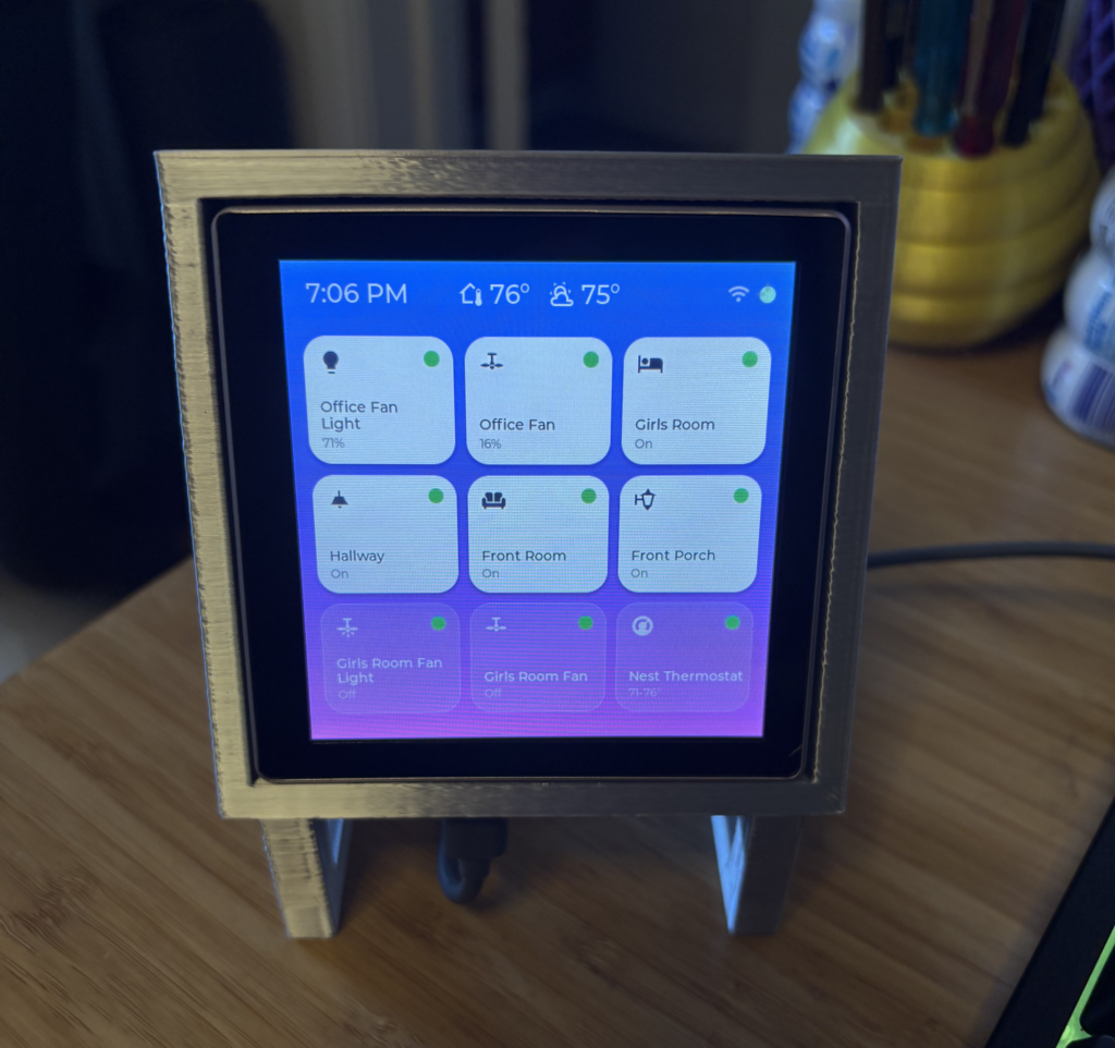 A small smart home control panel display in a gold 3D-printed frame showing nine device tiles including lights, fans, and a Nest thermostat, with the time reading 7:06 PM and indoor/outdoor temperatures of 76° and 75°.