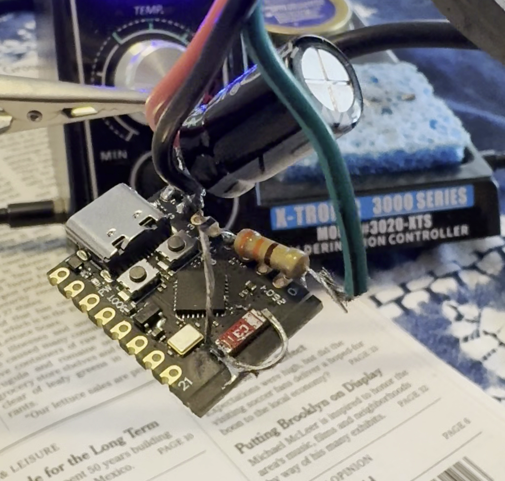 A small microcontroller PCB held in helping-hand clips with red, green, and black wires soldered to it, with a makeshift wifi antenna soldered to it.