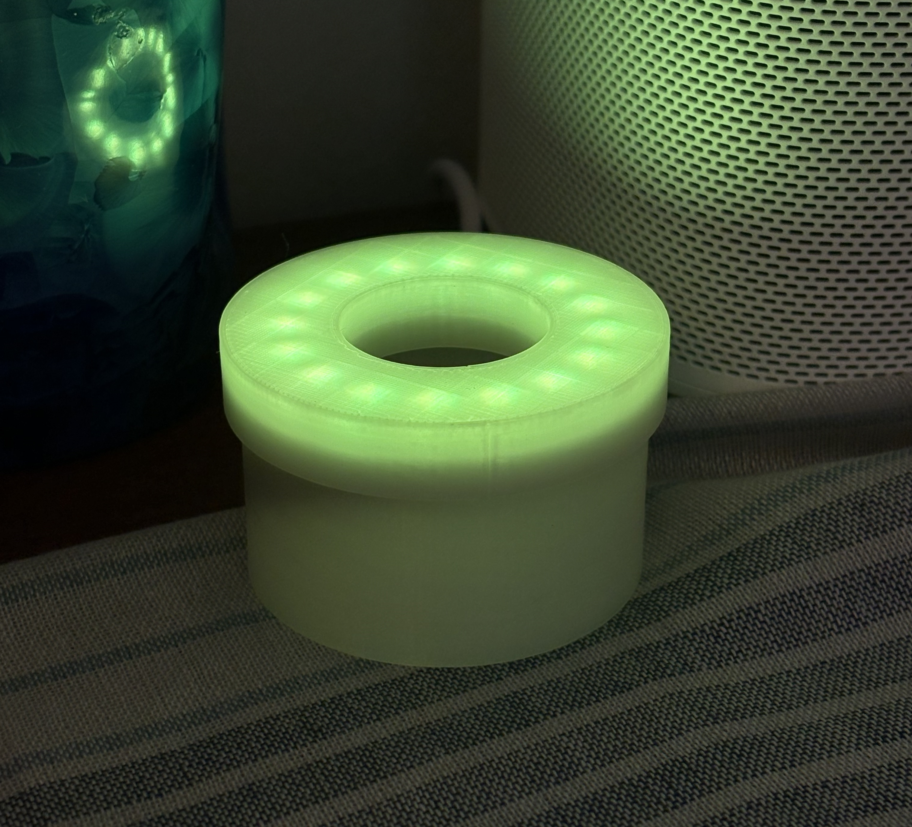 A glowing green 3D-printed cylindrical object with a hollow toroidal top containing LED lights, sitting on a striped fabric surface in a dimly lit room beside a smart speaker.