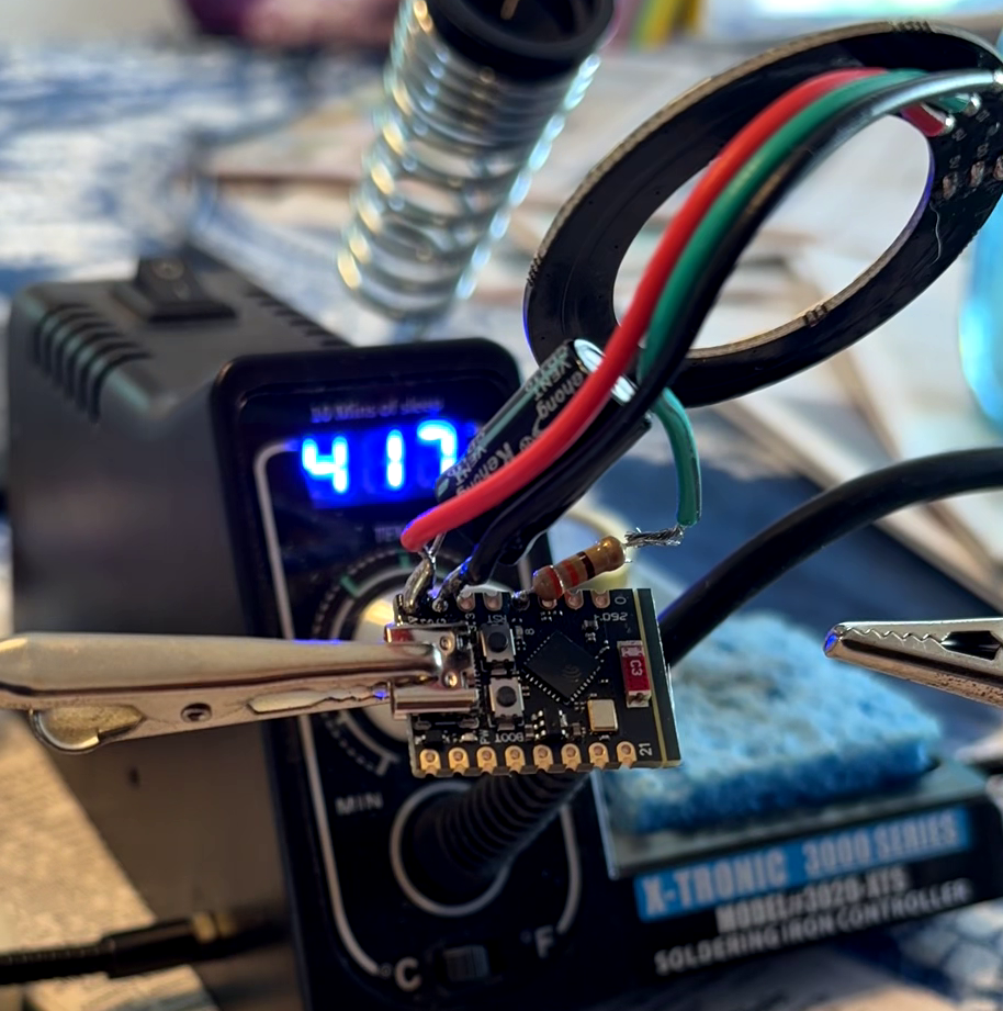 A small microcontroller PCB held in helping-hand clips with red, green, and black wires soldered to it, positioned in front of an X-Tronic 3020-XTS soldering iron controller displaying 417°C.