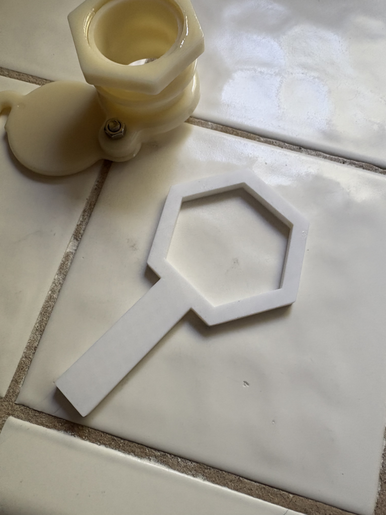 A hand holds a white plastic tool featuring a hexagonal nut shape over a white ceramic tiles in a kitchen. 