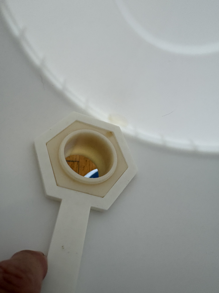A 3D-printed hexagonal wrench, fitted to a hexagonal nut on a honey gate inside a white 5 gallon bucket.