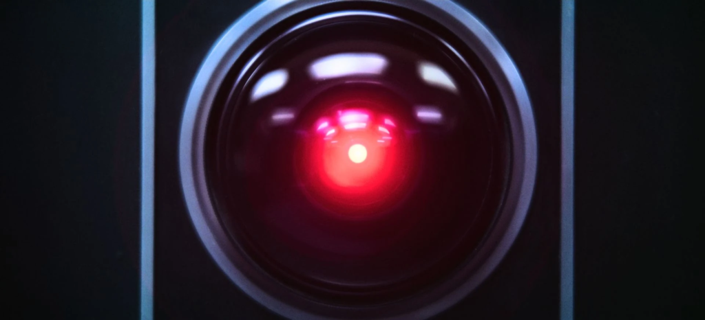 A close-up of HAL 9000's iconic camera eye from Stanley Kubrick's *2001: A Space Odyssey*, featuring a glowing red light at the center of a dark fisheye lens encased in a metallic ring.