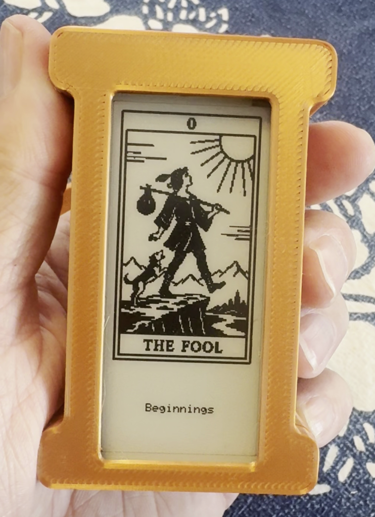 A hand holds a small e-ink display in a yellow 3D-printed case showing "The Fool" tarot card (numbered 0) with the word "Beginnings" beneath it, depicting the image of a figure with a bindle walking near a cliff's edge accompanied by a dog.