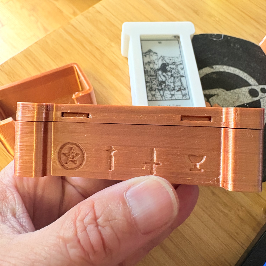 A hand holds a 3D-printed orange-brown rectangular box with embossed symbols including a pentacle, a wand, a sword, and a cup, with a white e-ink display device and wooden surface visible in the background.