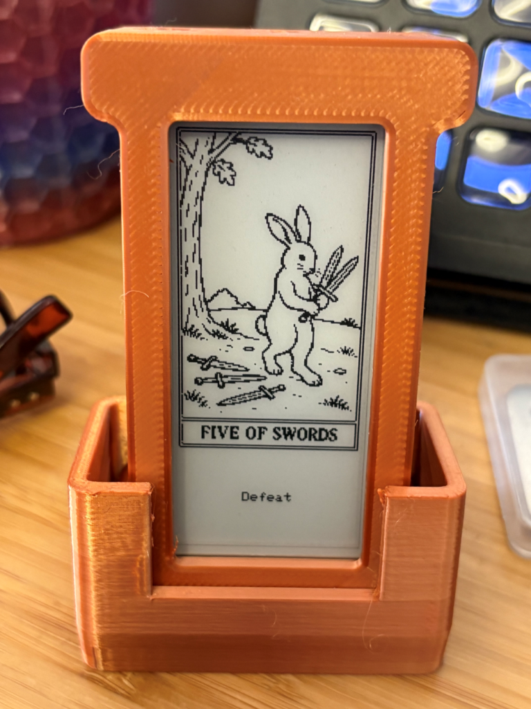 A 3D-printed orange stand holds a small e-ink display showing a pixel-art style tarot card — the Five of Swords — depicting a rabbit holding two swords with three more on the ground, labeled "Defeat" beneath the card border.