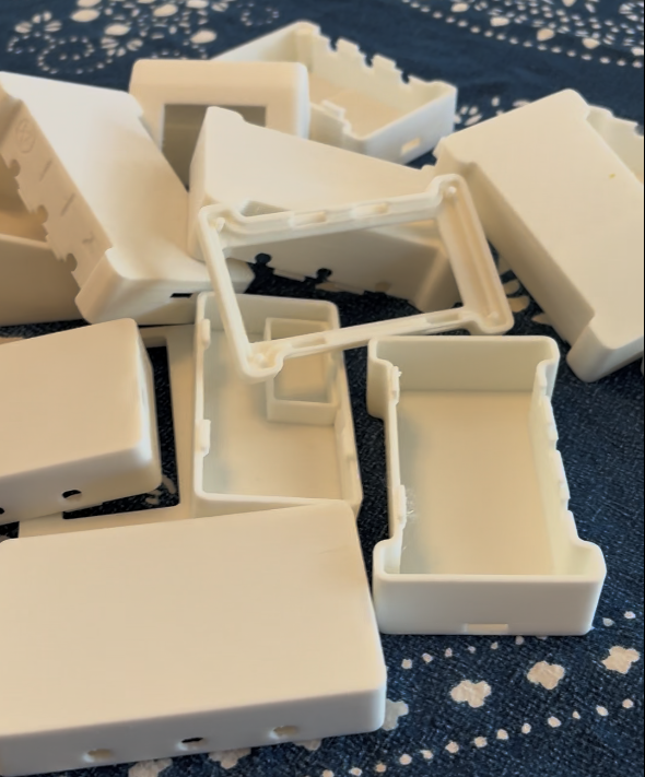 A pile of white 3D-printed plastic enclosure boxes with interlocking edges, scattered on a blue fabric surface, some still connected by print supports.